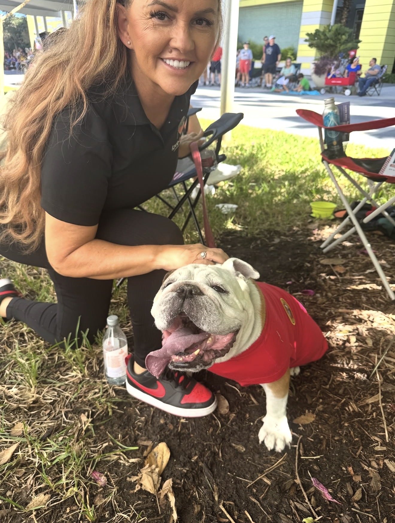A Love4Vets team member kneeling on the grass at a community event, smiling beside a bulldog in a red vest.